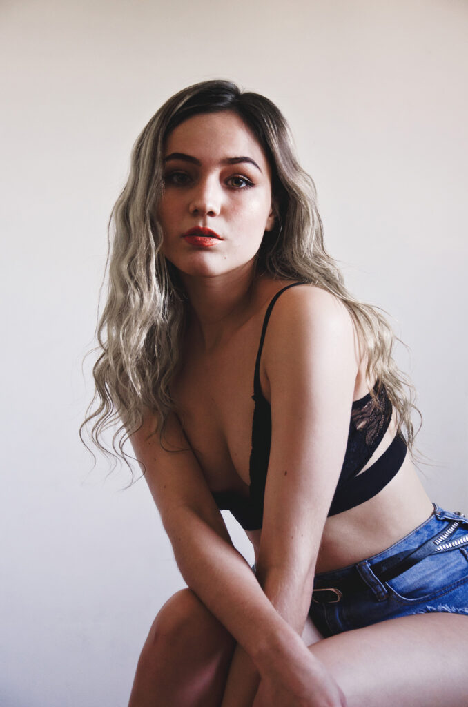Darian Rojas a model with long wavy blonde hair poses against a white backdrop. She wears a black lace bralette and blue denim shorts with a belt. Her gaze is directed towards the camera and she has a neutral expression.